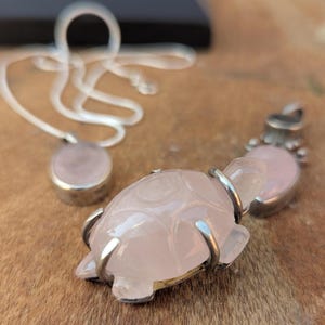 May include: A silver chain necklace with two pink rose quartz pendants. One pendant is a round stone, and the other is a carved turtle.