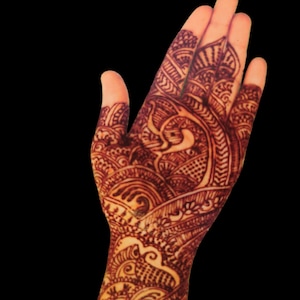 May include: Close-up of a hand with intricate henna designs. The henna is a deep reddish-brown color, applied in detailed patterns across the palm and fingers. The background is solid black, highlighting the henna art.