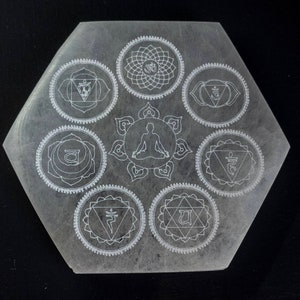 May include: A white hexagonal selenite charging plate with seven etched chakra symbols, including the root, sacral, solar plexus, heart, throat, third eye, and crown chakras. The plate also features a stylized figure sitting in meditation.