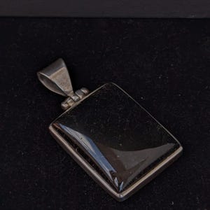 May include: A rectangular pendant with a polished black stone set in a silver-colored metal frame. The pendant has a loop at the top for a chain. The background is a dark, solid color.