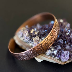 May include: A copper bracelet with an intricate design, resting on a bed of amethyst crystals.
