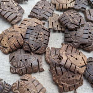 May include: A collection of brown coconut shell buttons with a leaf design. The buttons have two holes each and are arranged in a random pattern.