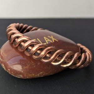 May include: A copper-colored bracelet with a twisted rope design rests on a reddish-brown stone. The stone has the word "RELAX" in gold lettering. The bracelet is made of a series of intertwined loops.