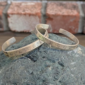 May include: Two silver bracelets with engraved Tibetan Buddhist symbols. The bracelets are stacked on top of each other on a gray rock.