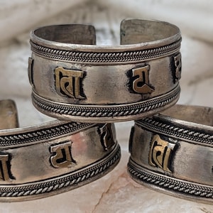 May include: Three silver bracelets with a braided design and gold lettering. The bracelets are stacked on top of each other.