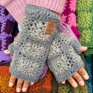 May include: Gray crocheted fingerless gloves with a brown leather tag that says "The Lazy Daisy". The gloves are made with a flower pattern and are shown against a background of colorful crocheted squares.