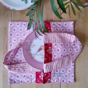 Può includere: Una tovaglietta quadrata in tessuto con motivo floreale rosa, una striscia centrale a fiori rossi e manici a quadretti vichy rosa chiaro. Un piatto rotondo con l'illustrazione di un uccellino poggia sulla tovaglietta, che reca l'etichetta Alice Design.