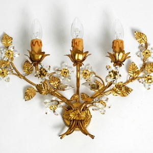 May include: A gold-toned metal wall sconce with a floral design. The sconce has three lights and is decorated with crystal accents.