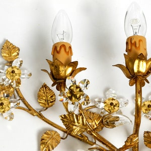 May include: Gold metal wall sconce with a floral design. The sconce features crystal flowers and leaves, and two candle-style light fixtures.