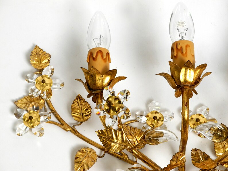 May include: Gold metal wall sconce with a floral design. The sconce features crystal flowers and leaves, and two candle-style light fixtures.