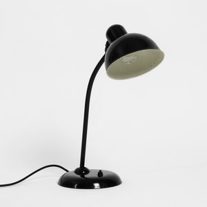 Puede incluir: Una l&aacute;mpara de escritorio negra con un brazo curvo y una pantalla redonda. La l&aacute;mpara tiene un interior blanco y un cable negro.