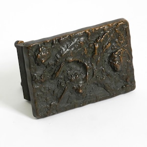 Puede incluir: Una escultura rectangular de color bronce con una superficie texturizada y un diseño en relieve. La escultura tiene un aspecto oscuro y envejecido, con detalles visibles de rostros y figuras. Está montada sobre una base negra, lo que sugiere que es un objeto decorativo.