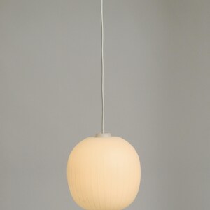 Puede incluir: L&aacute;mpara colgante redonda de color crema con detalles acanalados verticales. Cuelga de un fino cable blanco y tiene un dise&ntilde;o minimalista. La l&aacute;mpara tiene forma de esfera.