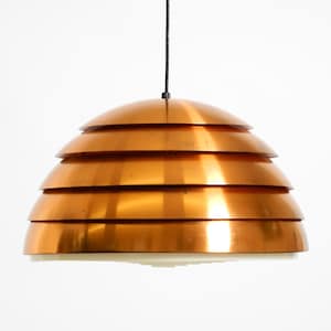 May include: A copper-colored pendant light fixture with a layered, tiered design. The light has a series of horizontal, curved metal shades, creating a unique, modern aesthetic. The fixture hangs from a black cord against a white background.
