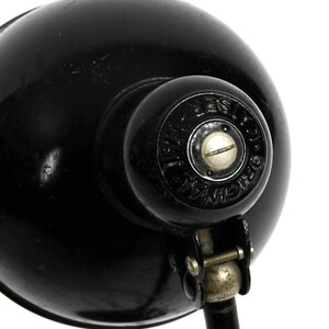 Puede incluir: L&aacute;mpara de escritorio de metal negro con una pantalla redonda y un brazo ajustable plateado. La l&aacute;mpara tiene una base de metal negro y una perilla de metal plateado en la pantalla. La pantalla tiene el texto "J.W. Ostberg" impreso en ella.