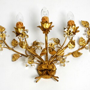 May include: A gold-toned metal wall sconce with a floral design. The sconce features three light bulbs and crystal accents.