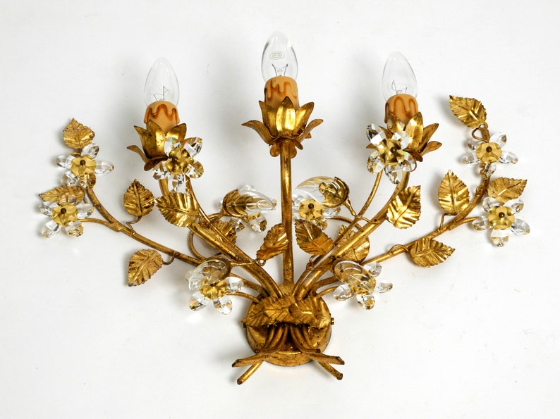 May include: A gold-toned metal wall sconce with a floral design. The sconce features three light bulbs and crystal accents.