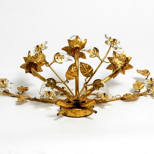 May include: Gold-toned metal ceiling light fixture with a floral design. The fixture features multiple branches with leaves and flowers, each with a clear glass crystal. The fixture is designed to be mounted on a ceiling.