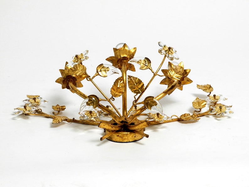 May include: Gold-toned metal ceiling light fixture with a floral design. The fixture features multiple branches with leaves and flowers, each with a clear glass crystal. The fixture is designed to be mounted on a ceiling.