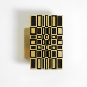 Puede incluir: Aplique de pared dorado con un patrón geométrico negro y dorado. El diseño presenta una serie de rectángulos y cuadrados dispuestos en forma de cuadrícula. El aplique está montado en una pared blanca, creando un contraste.