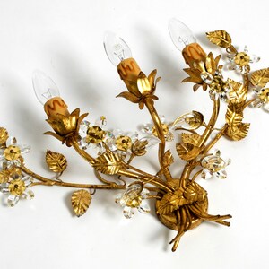 May include: Gold metal wall sconce with a floral design. The sconce has three light bulbs and is decorated with crystal accents.