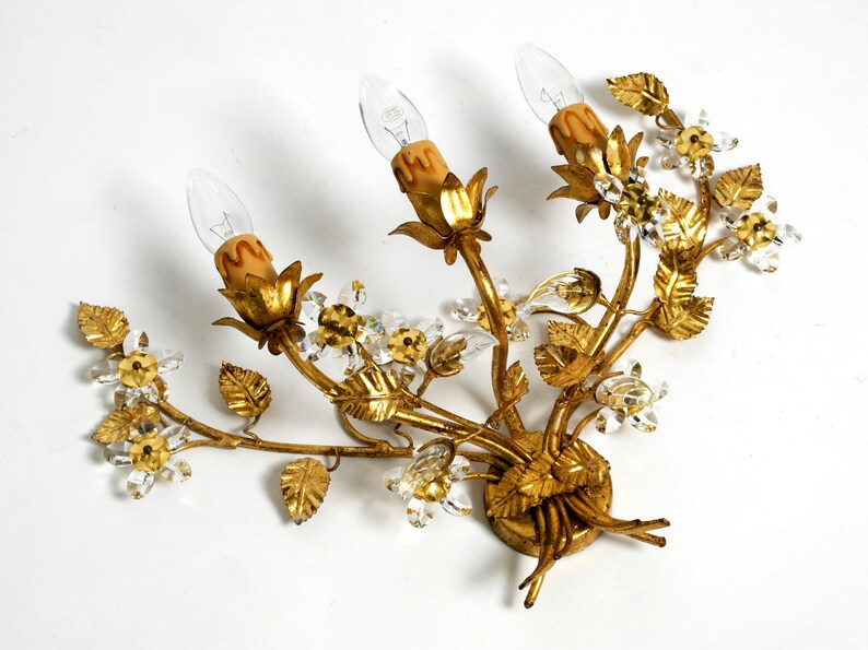 May include: Gold metal wall sconce with a floral design. The sconce has three light bulbs and is decorated with crystal accents.