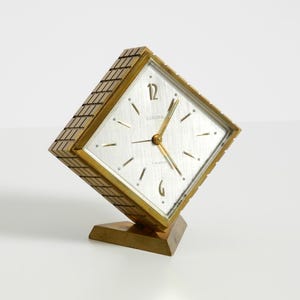 Könnte beinhalten: Eine Vintage-Quarzuhr in Goldoptik mit quadratischem Zifferblatt und einzigartigem, abgewinkeltem Design. Das Zifferblatt ist silberfarben mit schwarzen Stundenmarkierungen und Zeigern. Die Uhr ist mit "Europa" beschriftet und hat einen kleinen Sockel.