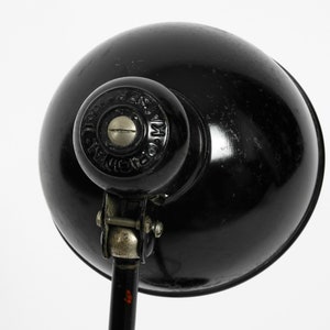 Puede incluir: L&aacute;mpara de escritorio de metal negro con una pantalla redonda y un brazo ajustable plateado. La l&aacute;mpara tiene un pomo negro con el texto "LICHT & M&Ouml;BEL" en blanco.