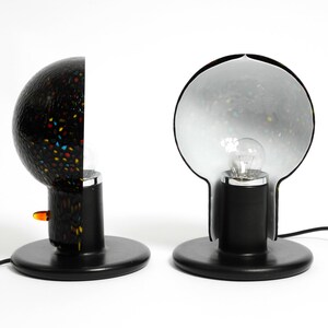 May include: Two black table lamps with a unique bulb shape. One lamp has a black and white speckled shade, while the other has a clear shade with a black and white speckled pattern.