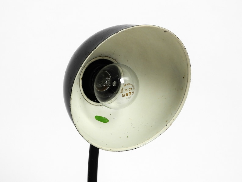 Puede incluir: L&aacute;mpara de escritorio negra y blanca con interior blanco y brazo de metal negro. La l&aacute;mpara tiene una bombilla de vidrio transparente con el texto "KESS 40 W" impreso en ella.