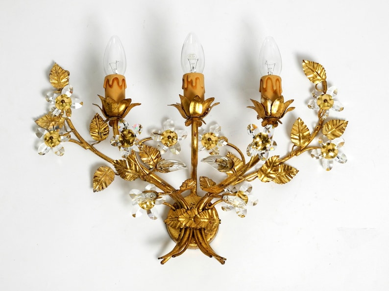May include: A gold-toned metal wall sconce with a floral design. The sconce has three lights and is decorated with crystal accents.