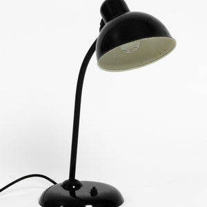 Puede incluir: Una l&aacute;mpara de escritorio negra con una pantalla interior blanca y un brazo flexible. La l&aacute;mpara tiene una base redonda y un cable.