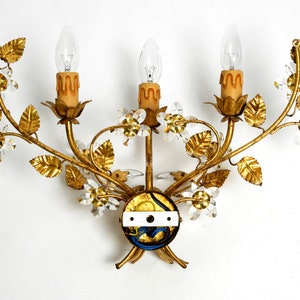 May include: Gold metal wall sconce with three candle-style light bulbs and crystal accents. The sconce is shaped like a floral branch with leaves and flowers.
