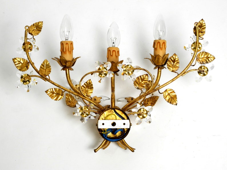 May include: Gold metal wall sconce with three candle-style light bulbs and crystal accents. The sconce is shaped like a floral branch with leaves and flowers.