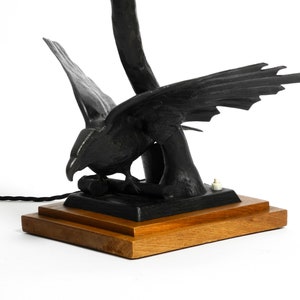 May include: Black metal table lamp with a bird sculpture base. The bird has its wings spread and is perched on a branch. The lamp has a wooden base.