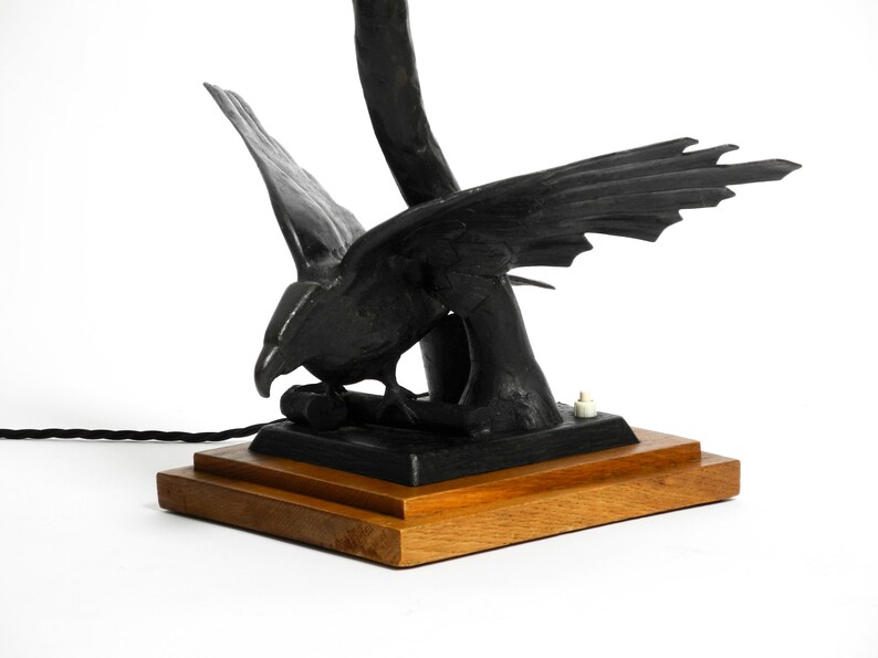 May include: Black metal table lamp with a bird sculpture base. The bird has its wings spread and is perched on a branch. The lamp has a wooden base.
