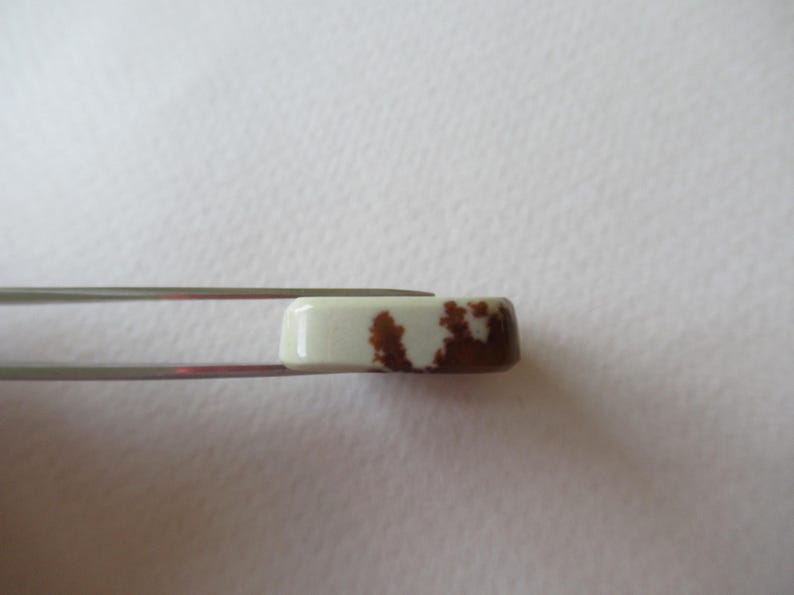 Pode incluir: Um close-up de um caboch&atilde;o retangular de pedra branca e marrom segurado por uma pin&ccedil;a. A pedra tem um padr&atilde;o manchado de marrom e branco.