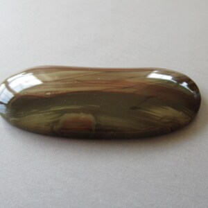 May include: A polished, oval-shaped gemstone with a glossy surface. The stone features a blend of brown, reddish-brown, and olive-green hues, with visible streaks and patterns. The gemstone is set against a neutral background.