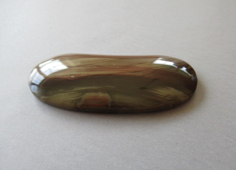 May include: A polished, oval-shaped gemstone with a glossy surface. The stone features a blend of brown, reddish-brown, and olive-green hues, with visible streaks and patterns. The gemstone is set against a neutral background.