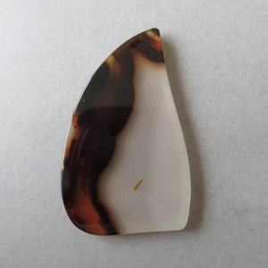 May include: A polished, teardrop-shaped gemstone with a striking pattern. The stone features a blend of colors, including white, brown, and orange, creating a unique, natural design. The gemstone is smooth and appears to be translucent.