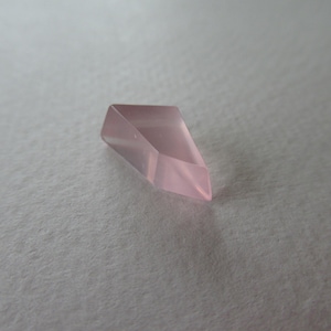 May include: A faceted, pale pink crystal with a pointed end. The crystal is a geometric shape, with multiple flat sides that reflect light. The background is a soft, textured, off-white surface.