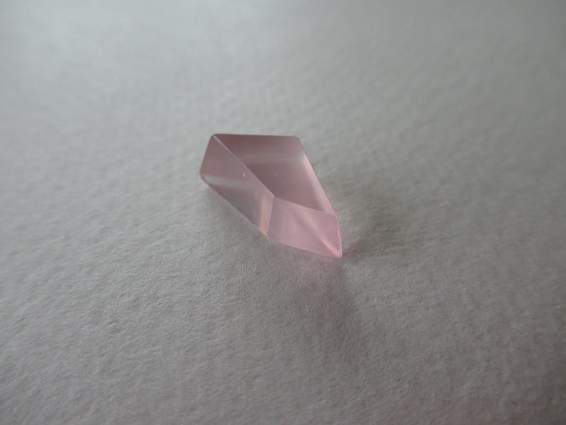 May include: A faceted, pale pink crystal with a pointed end. The crystal is a geometric shape, with multiple flat sides that reflect light. The background is a soft, textured, off-white surface.