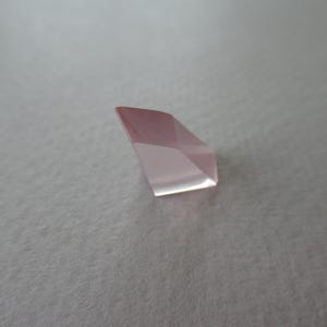 May include: A faceted, pale pink gemstone with a geometric shape. The gem has multiple flat surfaces that reflect light, creating a sparkling effect. The stone is set against a neutral background.