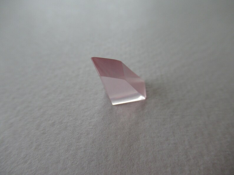 May include: A faceted, pale pink gemstone with a geometric shape. The gem has multiple flat surfaces that reflect light, creating a sparkling effect. The stone is set against a neutral background.