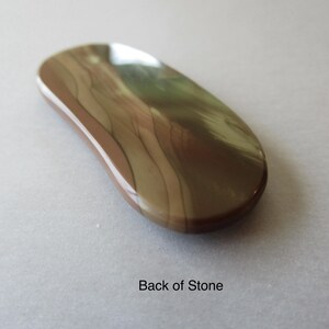May include: A polished, oval-shaped stone with a smooth, glossy surface. The stone features a blend of brown, tan, and cream colors with a wavy pattern. The text "Back of Stone" is visible.