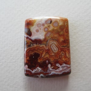 May include: A rectangular gemstone with rounded edges, showcasing a natural pattern of swirling colors. The stone features shades of brown, red, white, and cream, creating a unique, organic design. The polished surface reflects light, highlighting the intricate details.