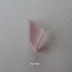 May include: A pale pink, geometric gemstone, viewed from above. The stone has a smooth, polished surface and a translucent quality. The shape is a modified triangle or kite, with a slightly rounded edge. The background is a neutral, off-white colour.