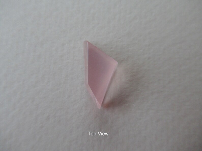 May include: A pale pink, geometric gemstone, viewed from above. The stone has a smooth, polished surface and a translucent quality. The shape is a modified triangle or kite, with a slightly rounded edge. The background is a neutral, off-white colour.