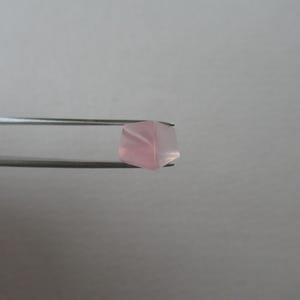 May include: A light pink, faceted gemstone is held by silver tweezers. The gemstone has a translucent appearance, with delicate internal patterns. The background is a plain, light grey.