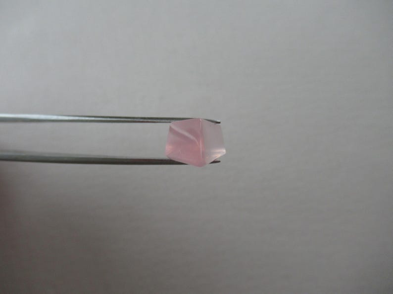 May include: A light pink, faceted gemstone is held by silver tweezers. The gemstone has a translucent appearance, with delicate internal patterns. The background is a plain, light grey.